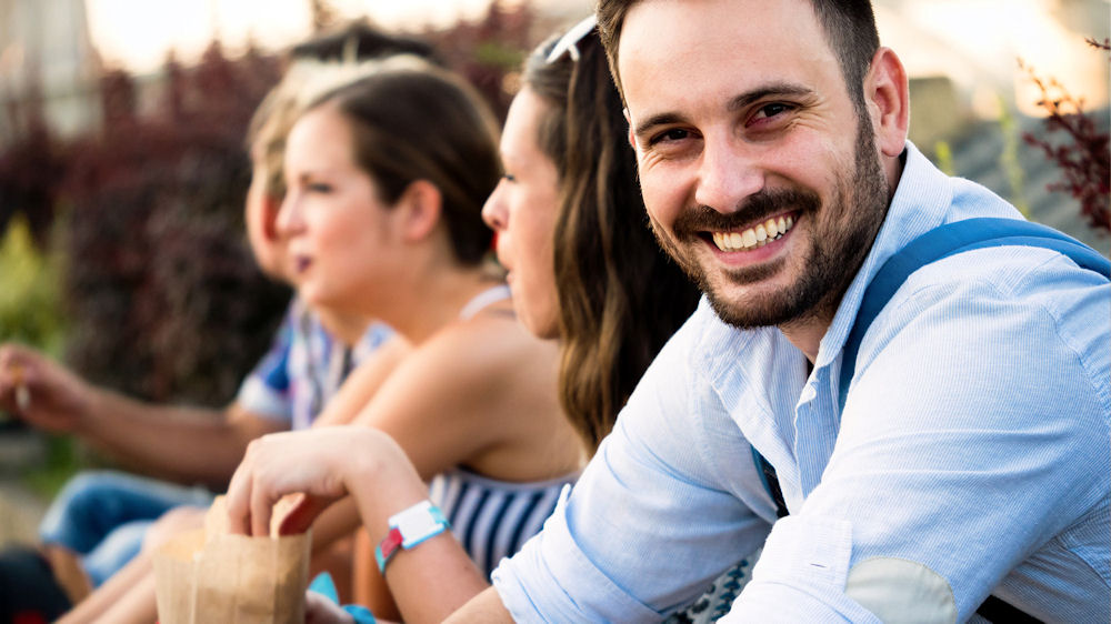 Smiling man with beard seated next to group of people on dock.